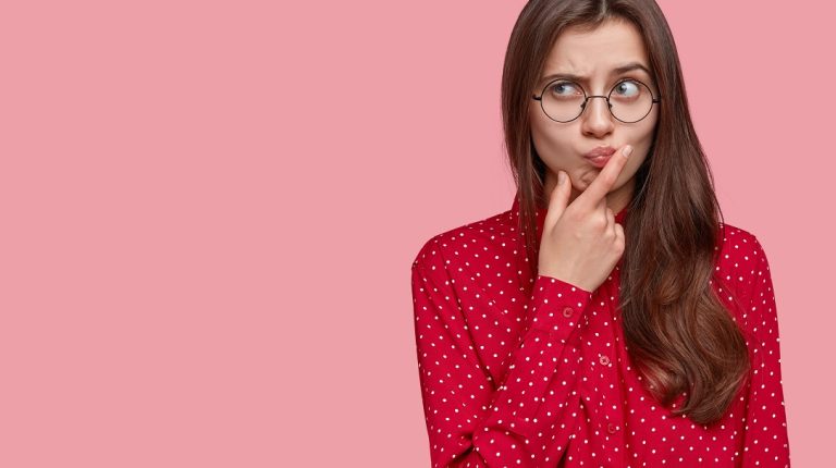 Young attractive woman with thoughtful expression, thinks about question, feels doubt, raises eyebrows, holds chin, dressed in formal clothes, isolated over pink background with free space on left
