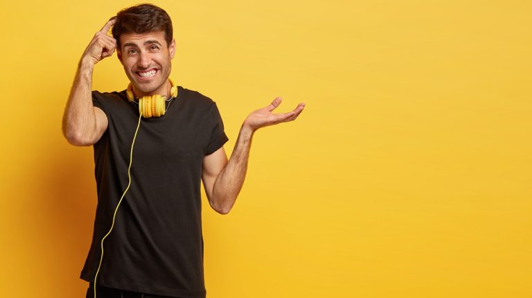 Hesitant unaware man scratches head, makes difficult decision, raises palm with hesitation, wears casual black t shirt, headphones on neck for listening music, isolated on yellow wall, empty space